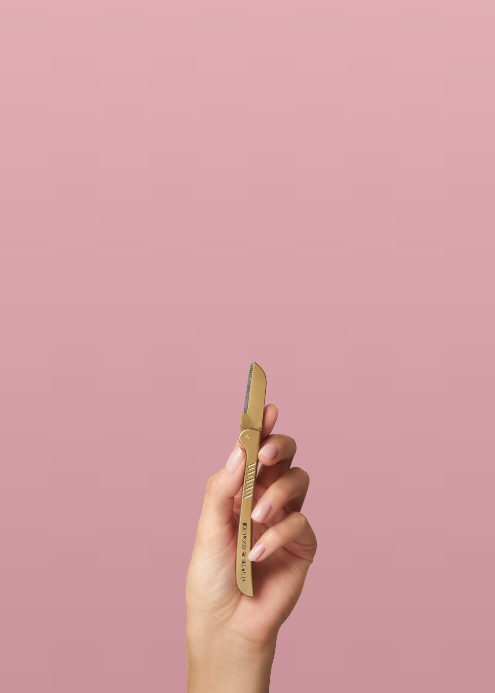 Hand holding a gold eyebrow razor against a pink background